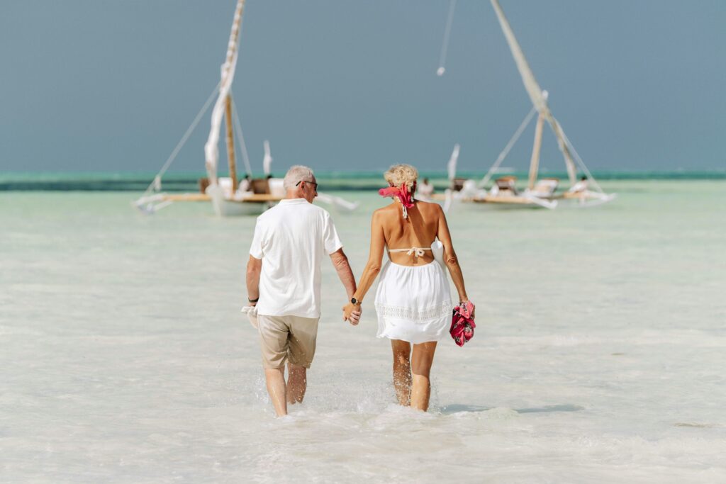 An elderly couple holds hands while walking on the beach, with dhows in the background in Zanzibar.